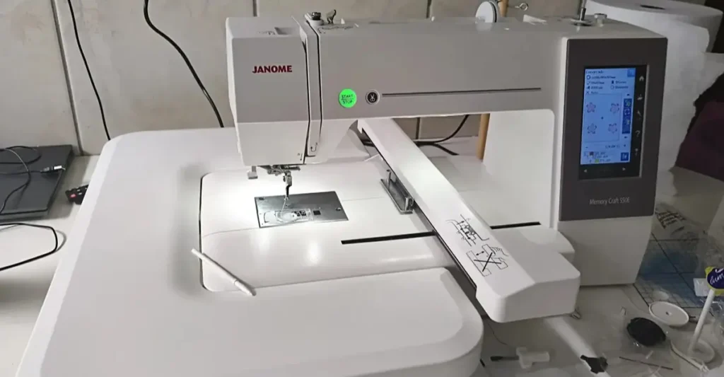 Máquina de bordar Janome MC 550e ligada na bancada do ateliê, com a mesa extensora branca acoplada, tela touch acesa e a caneta stylus apoiada, pronta para o teste prático.
