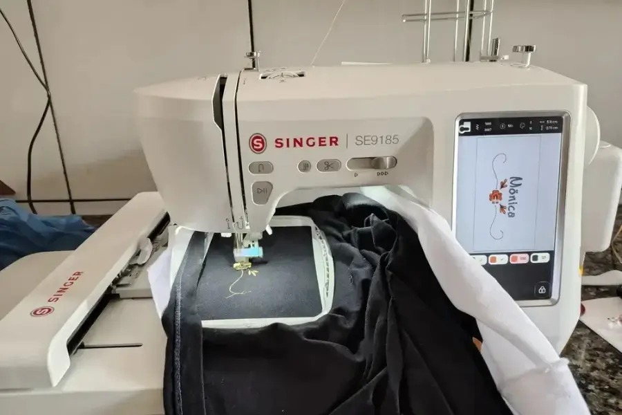 Máquina Singer SE9185 bordando um detalhe amarelo e o nome Mônica em um tecido preto preso ao bastidor.